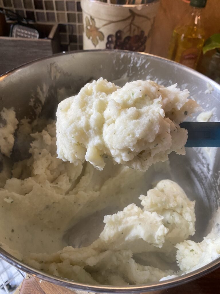 A spatula of creamy mashed potatoes over a metal bowl of the same
