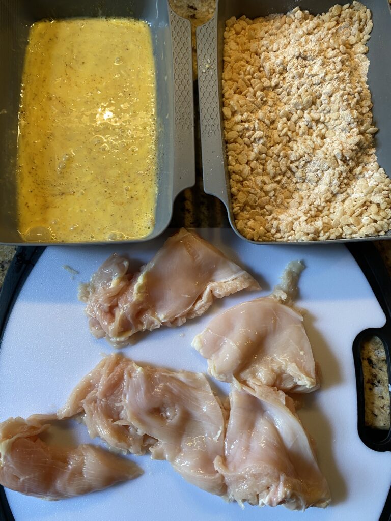 A dish of egg wash, a dish of Rice Crispies, and a cutting board with raw chicken on it