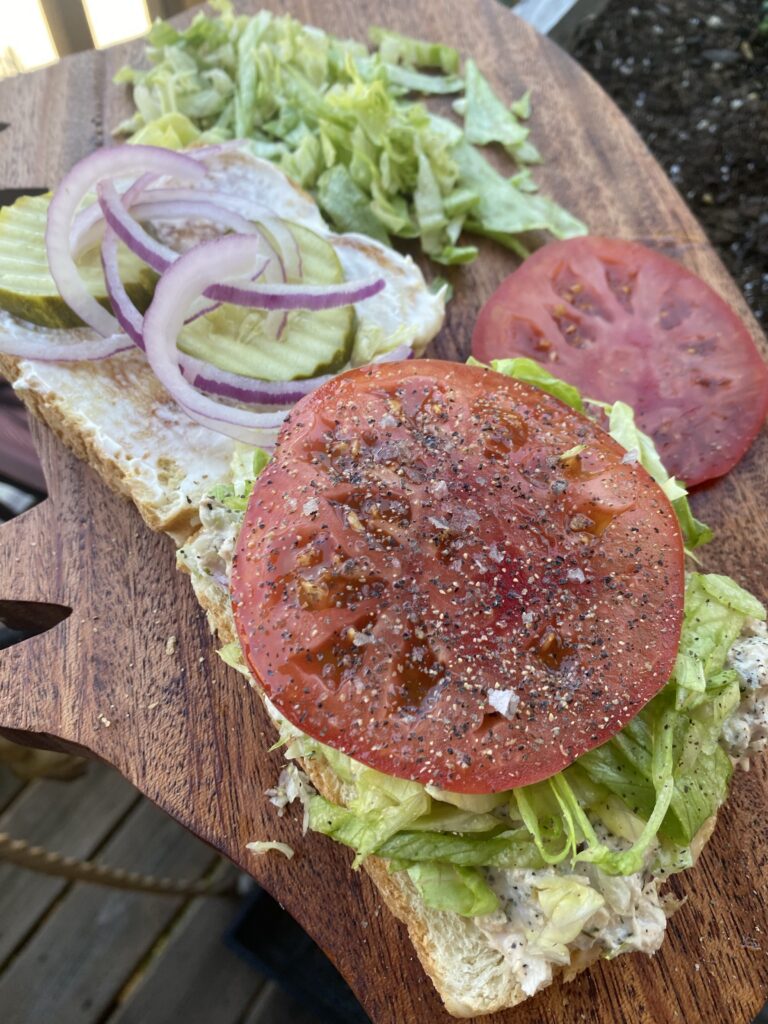 Picture of a garden fresh tomato with seal salt and black pepper on top of an open tuna salad sandwich