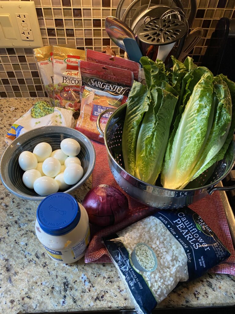 The ingredients for the seven layer salad, including the lettuce, hard boiled eggs, mayo, cheese, peas, and cauliflower pears