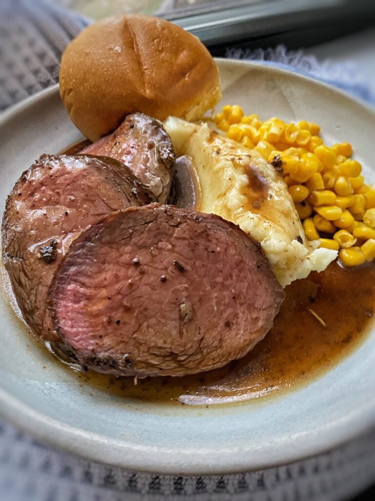 Slices of chuck tender roast with gravy, mashed potatoes, corn, and a roll