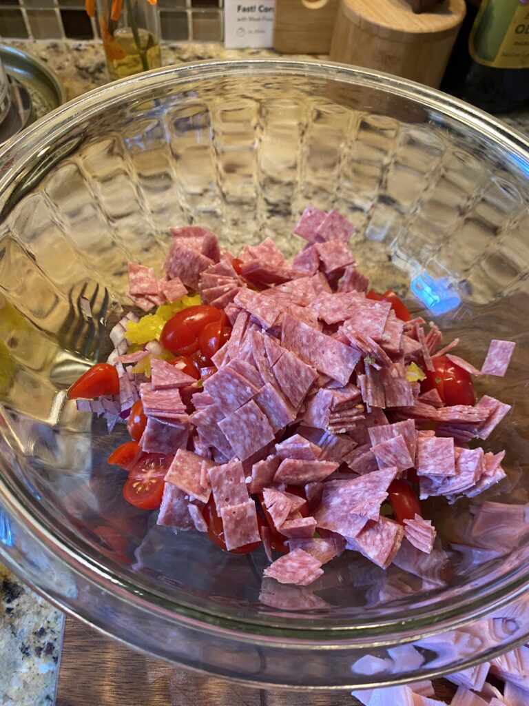 A glass bowl with chopped deli meat, grape tomatoes, and diced onions and banana peppers