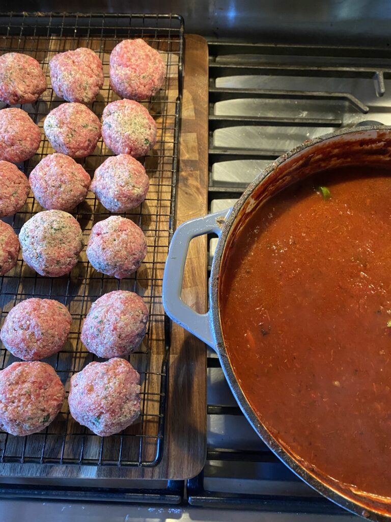 Uncooked meatballs on a metal grate next to a big pot of Sunday sauce
