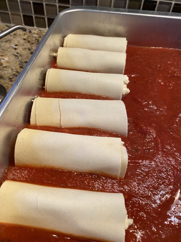 A row of manicotti noodles wrapped around ricotta cheese in marinara sauce 