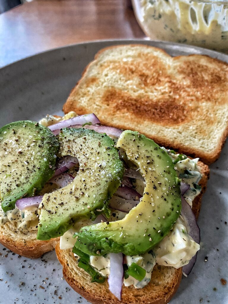 An open egg salad sandwich with avocado, onion, and seasonings