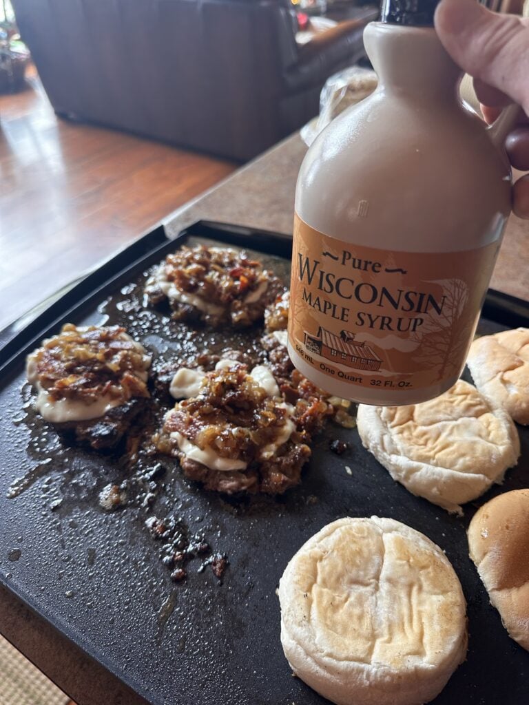 A bottle of syrup held over burgers cooking on a griddle