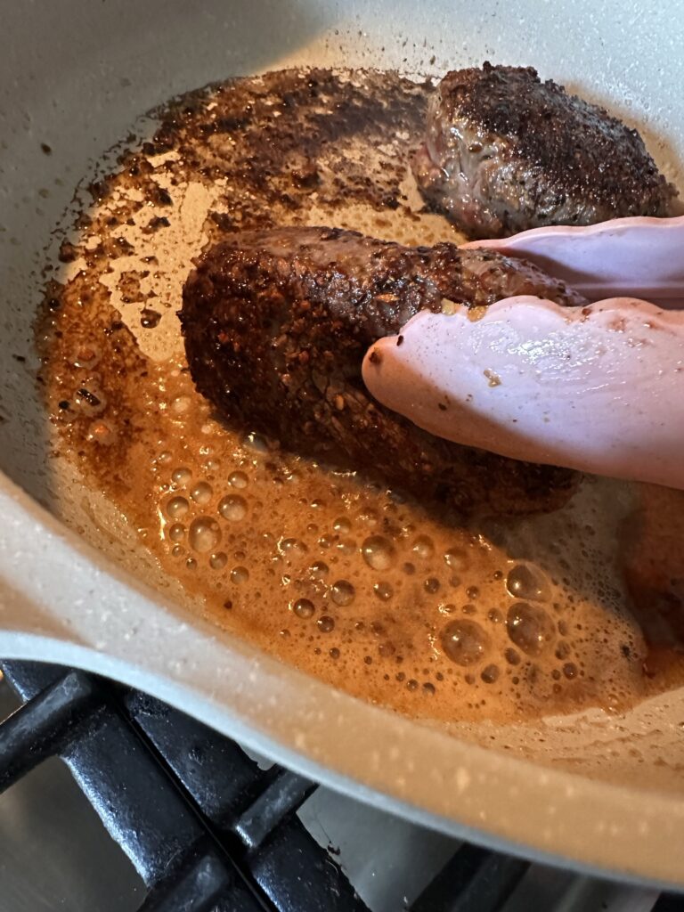 A steak cooking in a pan being flipped with tongs
