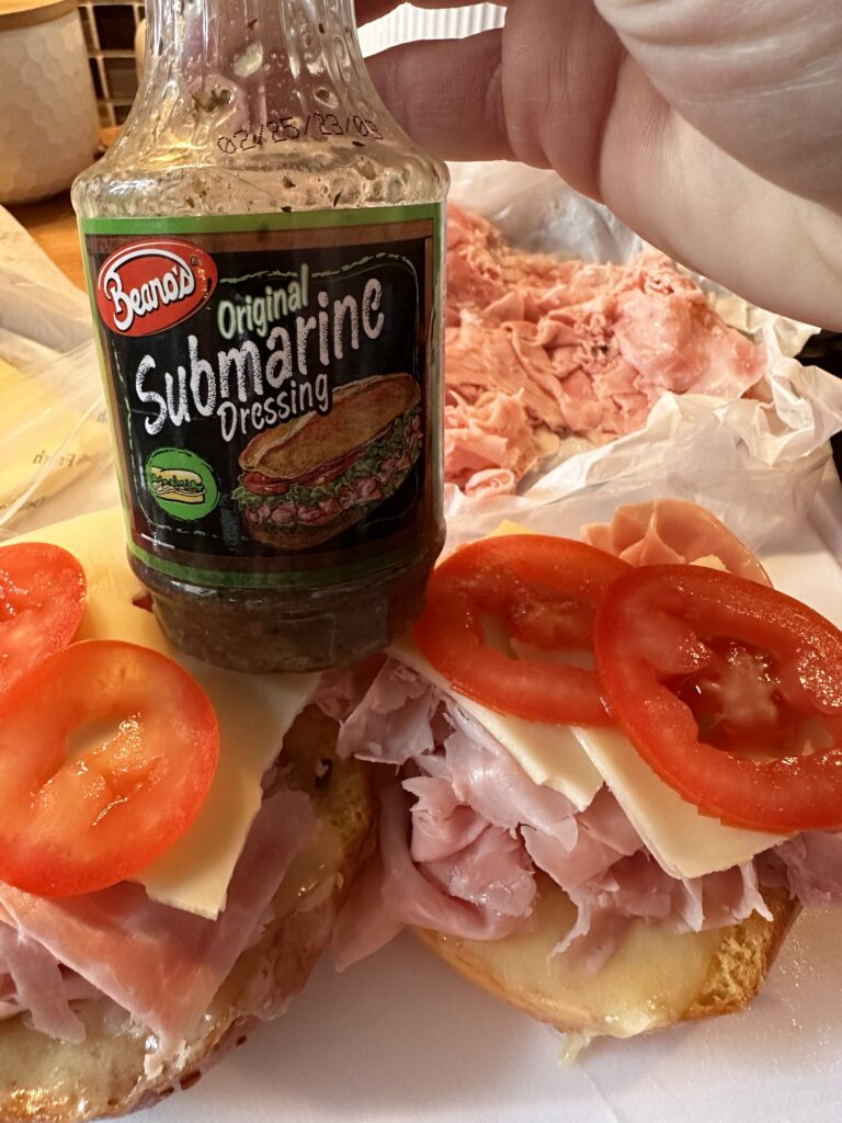 A bottle of Beano's original submarine dressing above hamburger buns topped with shaved ham, tomato slices, and cheese