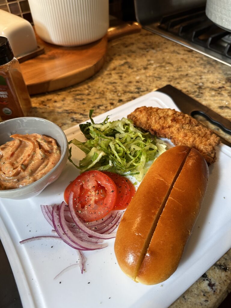 The components of a bird dog sandwich, including lettuce, onion, a chicken tender, and a hot dog bun 