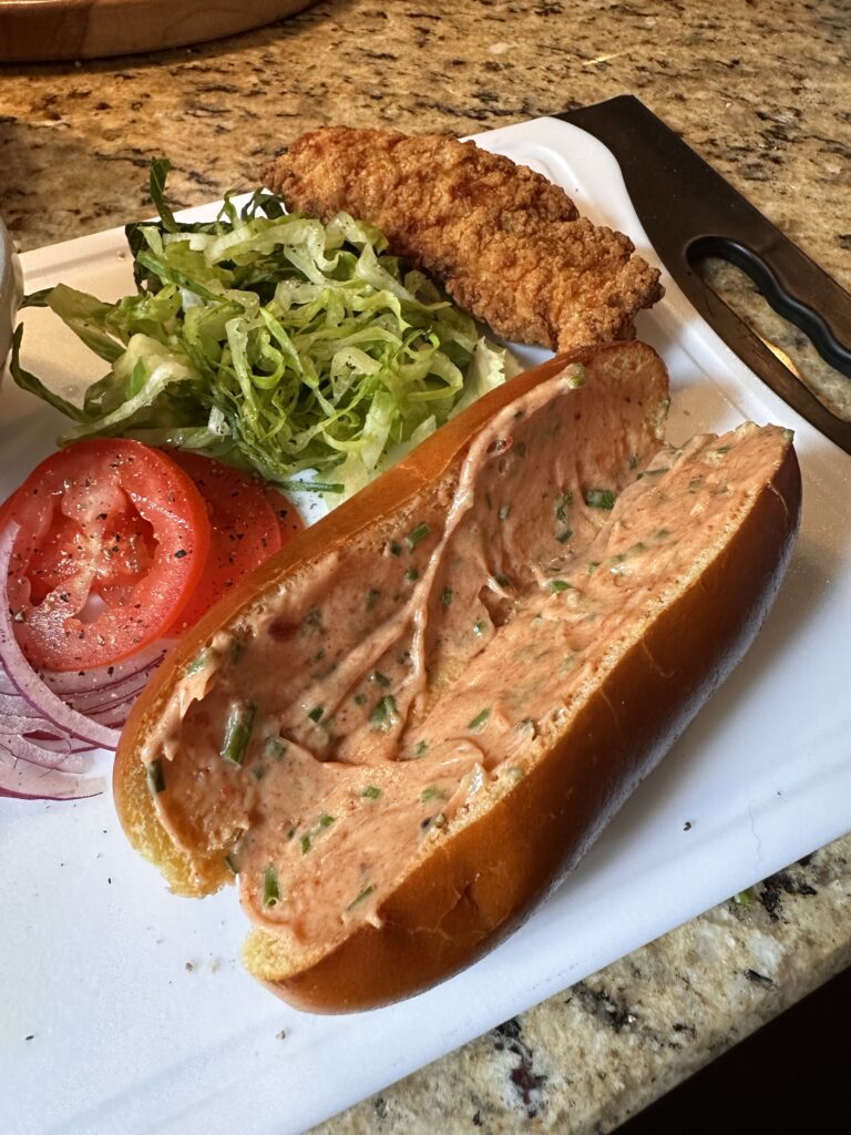 The components of a bird dog sandwich, including lettuce, onion, a chicken tender, and a hot dog bun with sauce spread in it