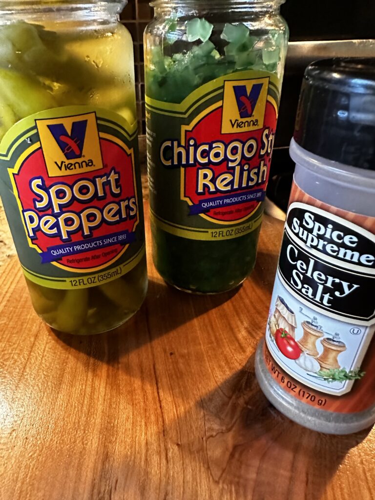 A jar of sport peppers, a jar of relish, and a shaker of celery salt