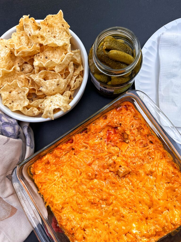 A glass dish of pickley buffalo chicken dip next to a bowl of corn chips
