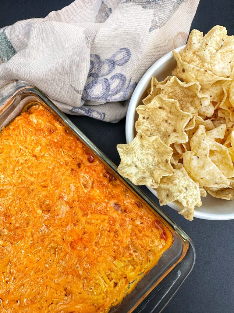 The pickley buffalo chicken dip in a glass dish next to a bowl of corn chips