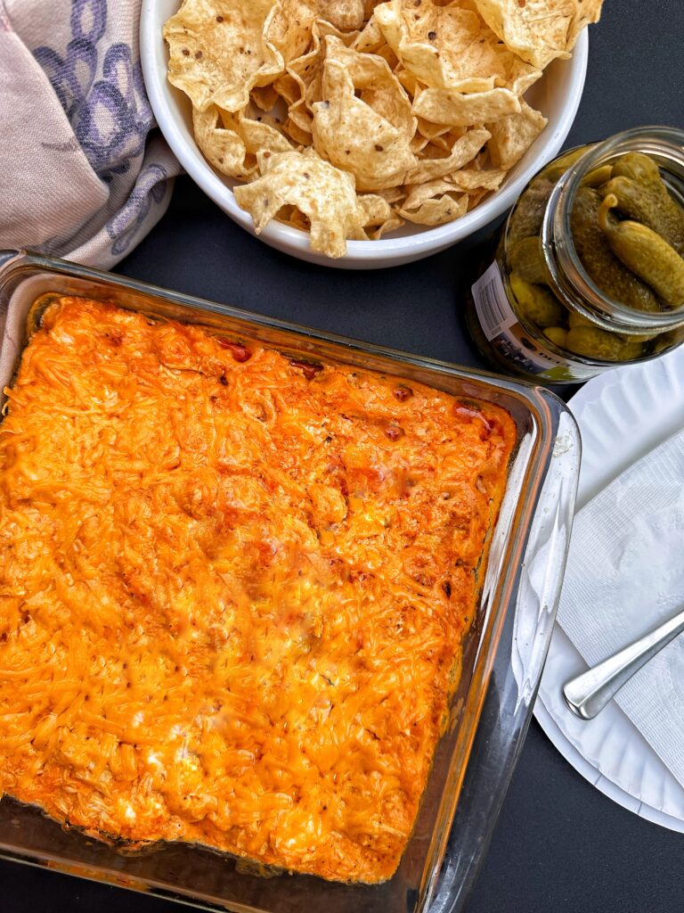 The finished dip next to a bowl of corn chips and a jar of pickles