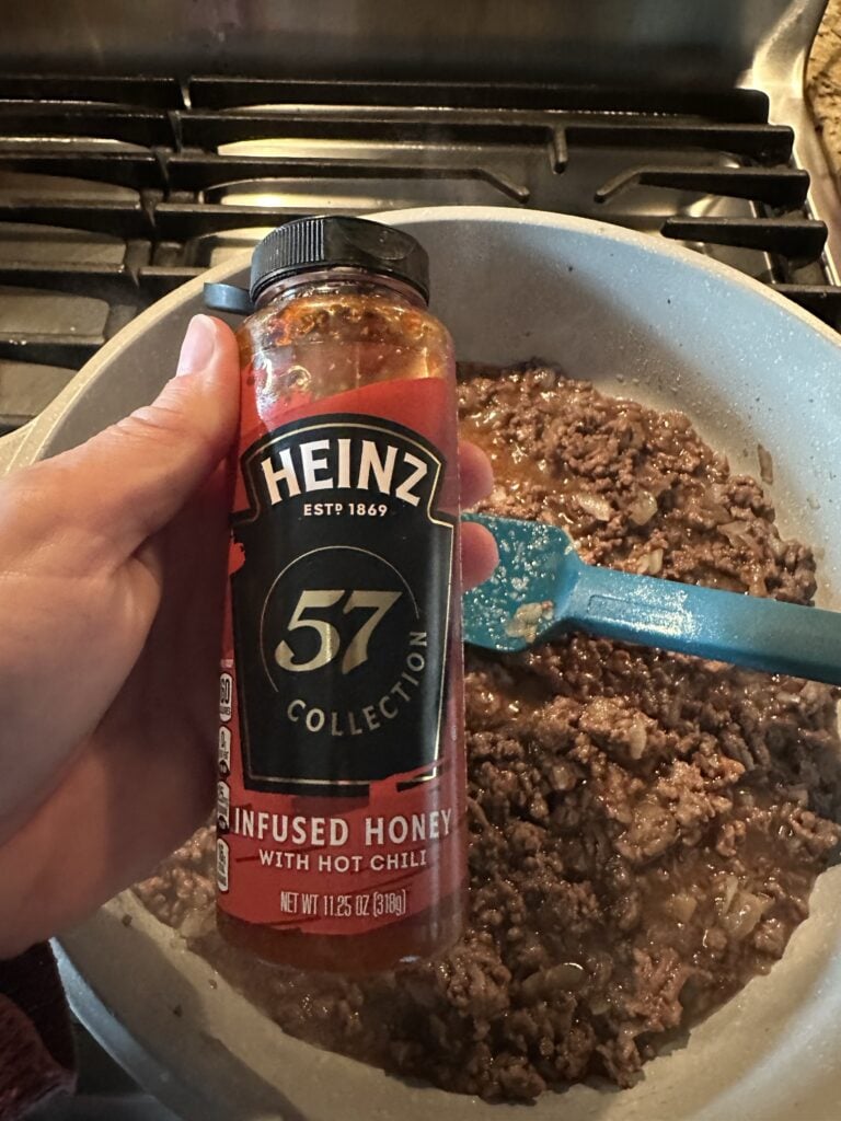 A bottle of Heinz infused honey held above a pot of cooking ground beef