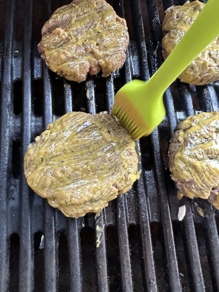 Burger patties on the grill being brushed with mustard