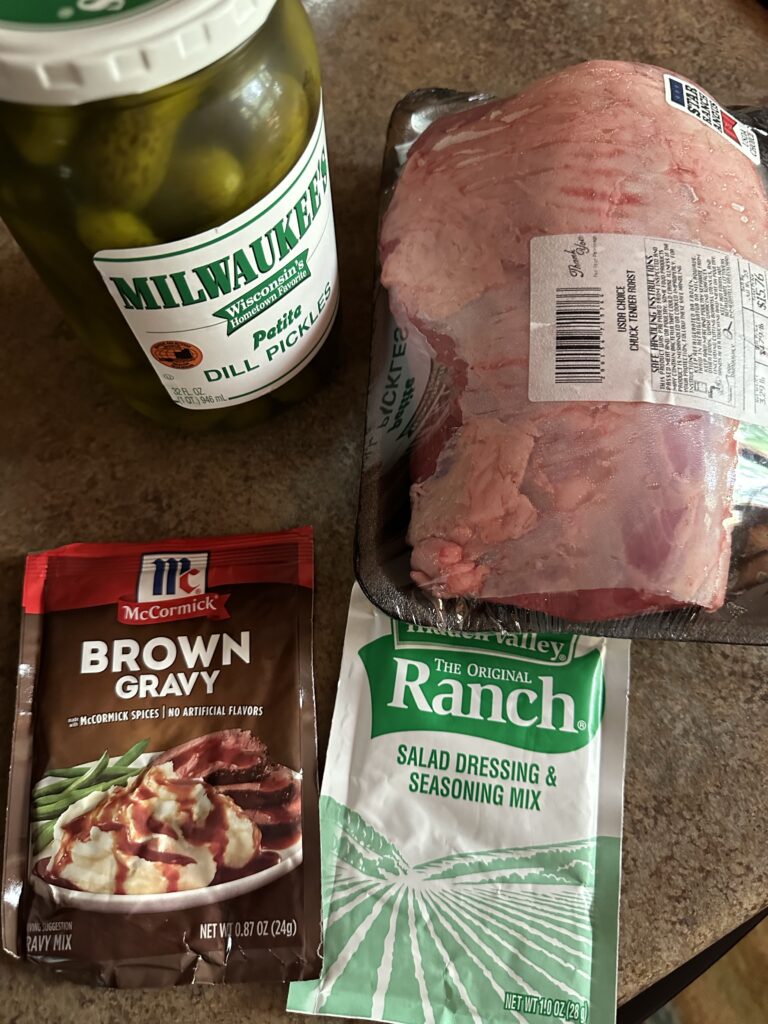 The ingredients for the recipe, including a jar of pickles, a package of beef chuck roast, a brown gravy packet, and a packet of ranch dressing mix