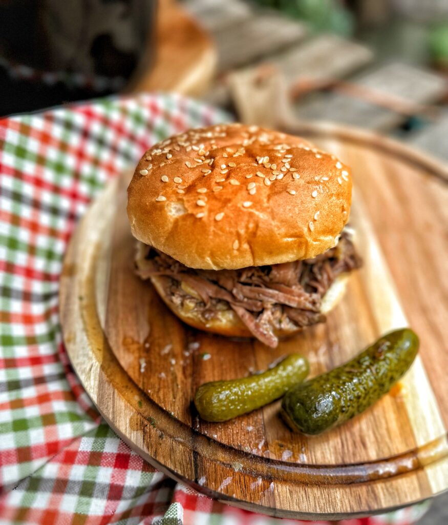 A pickle pot roast sandwich on a cutting board next to two pickles