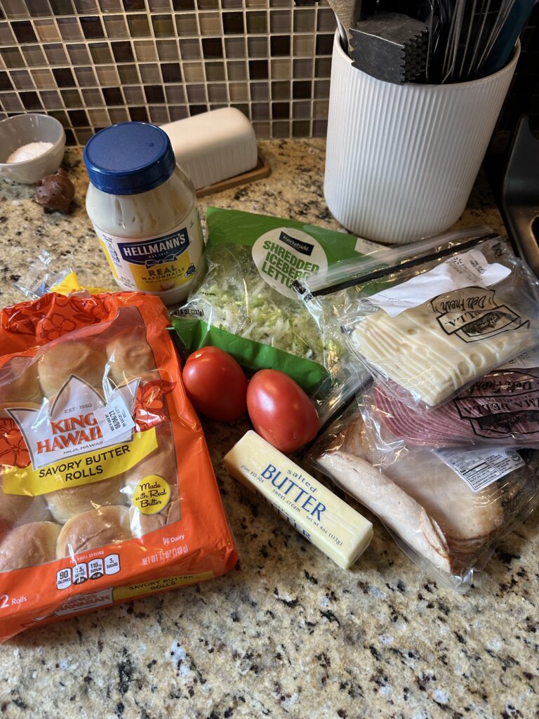 The ingredients for the boat bite sandwiches including King's Hawaiian rolls, mayo, shredded lettuce, sliced cheese, deli meats, butter, and tomatoes