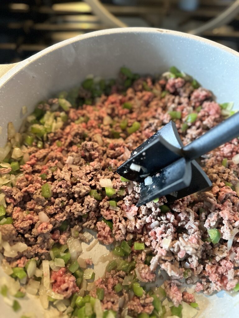 Ground beef and diced peppers and onions being broken up with a meat smasher while cooking in a pan