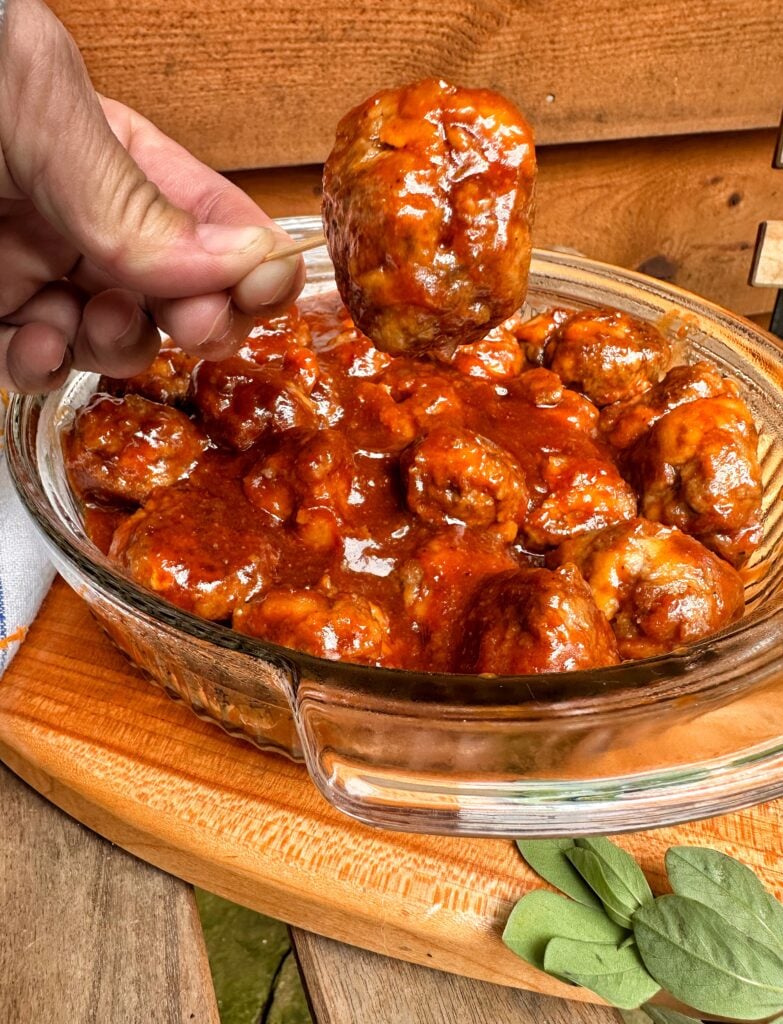 A Beef BBQ meatball held on a toothpick above a bowl of more meatballs