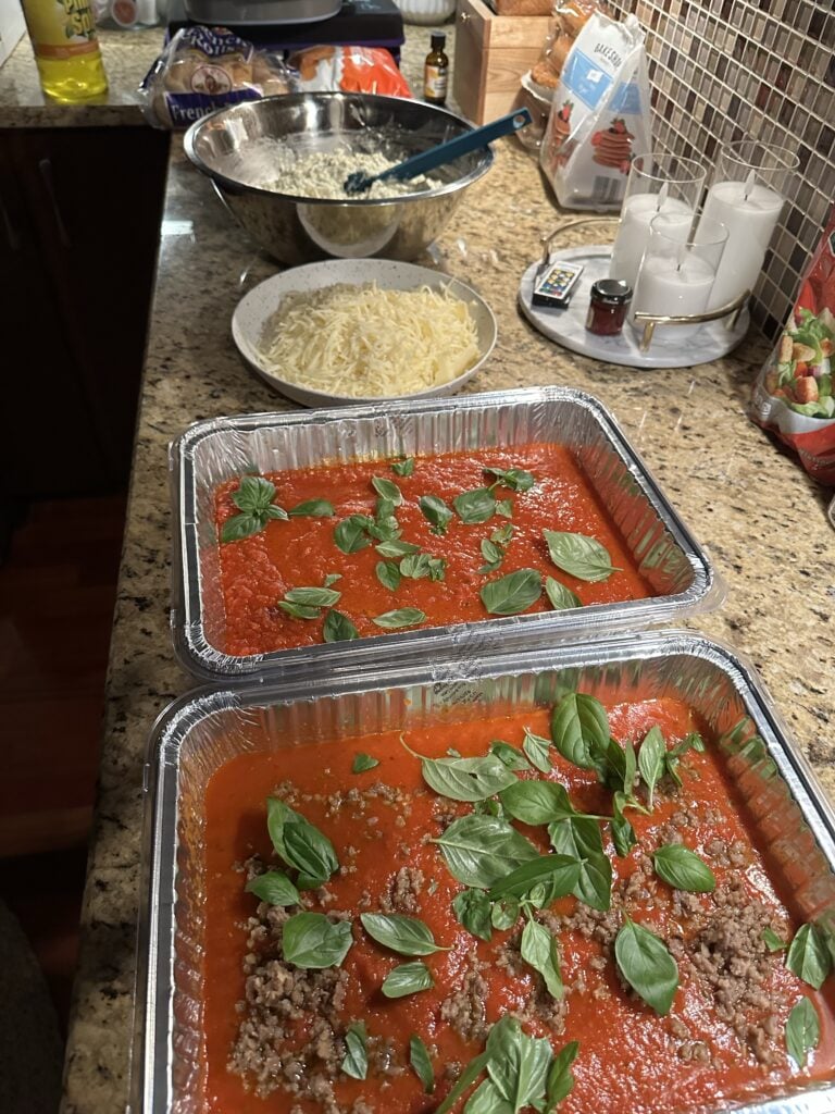 A station set up on the kitchen counter with everything needed to prepare the lasagna, including a bowl of cheese mixture, a bowl of shredded cheese, and aluminum tins with tomato sauce and basil, one with sausage and one without