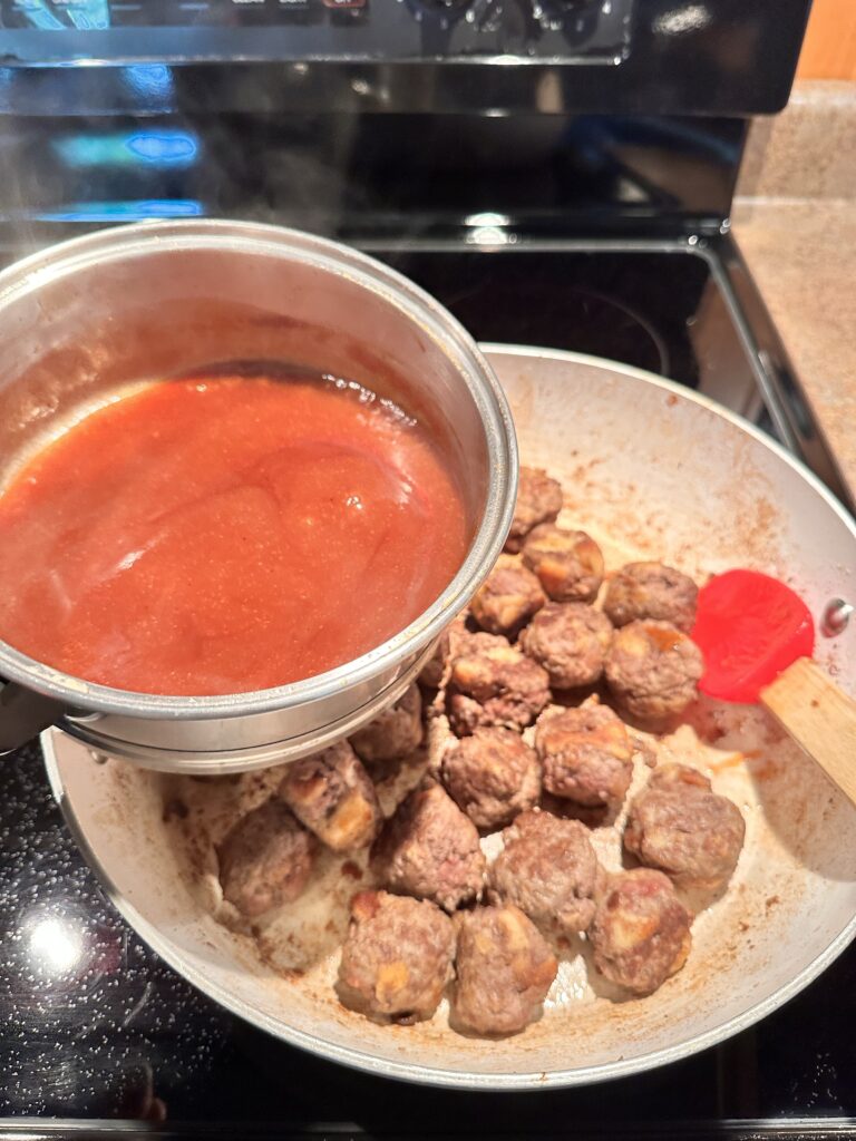 Meatballs cooking in a pan, about to be covered in sauce from a small pot