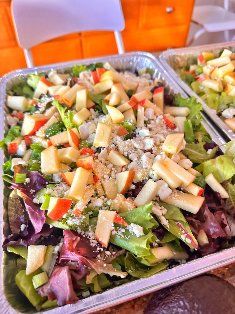 The fall salad in an aluminum baking dish topped with sliced apples and goat cheese crumble