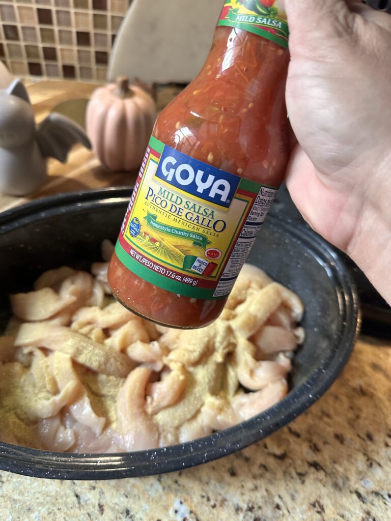 A bottle of Goya Pico De Gallo held in front of shredded raw chicken in a cooking dish