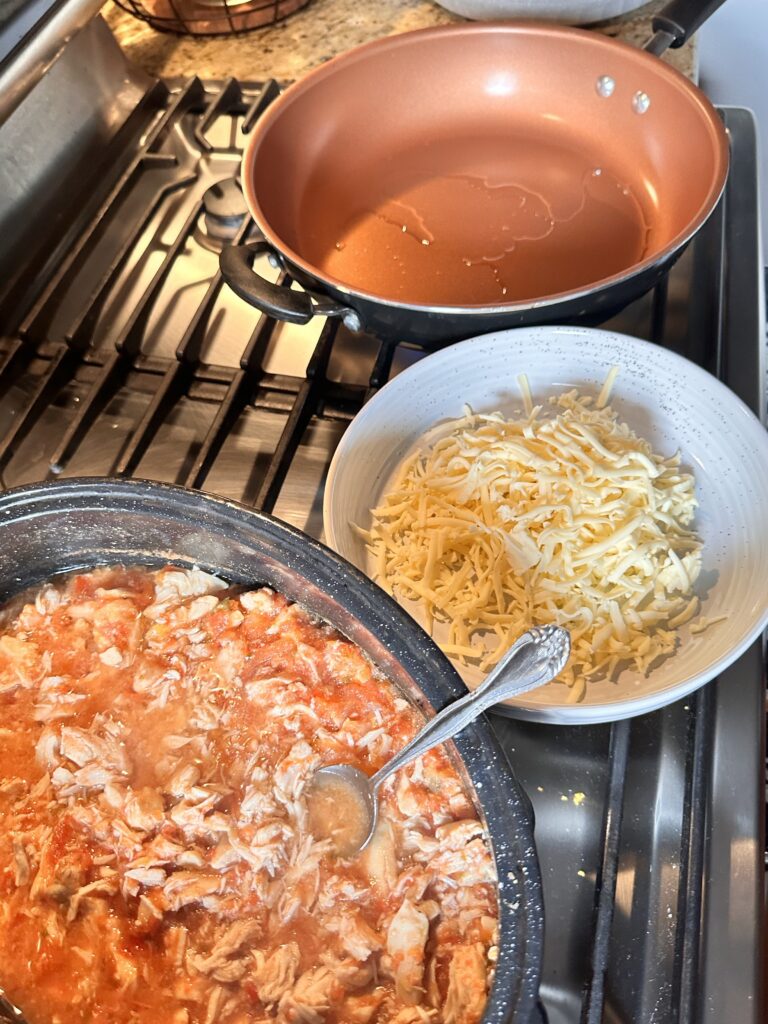 Shredded chicken in red sauce next to a bowl of shredded cheese and a pan with oil in it