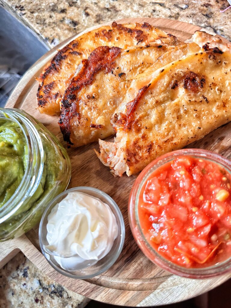 A close up of the finished crispy chicken tacos next to glass containers of guacamole, sour cream, and salsa