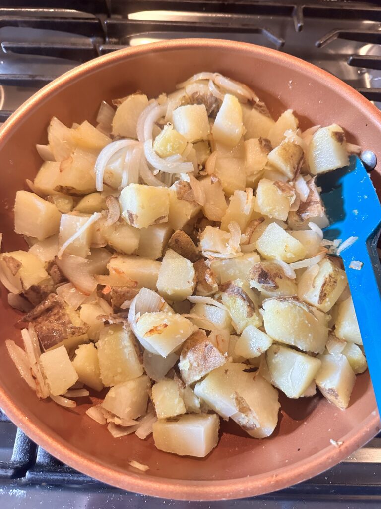 Cubed potatoes and sliced onions cooking in a pan with a blue spatula.