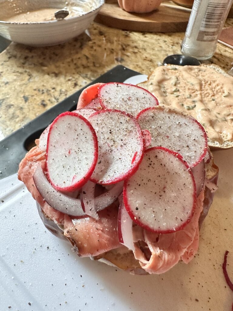The roast beef sandwich with the top half of the bun, which is spread with sauce, removed and the roast beef is topped with sliced onions and radish.