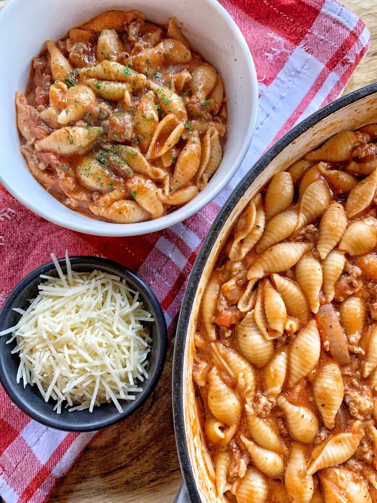 A bowl of beefy tomato cheese shells next to a pot of the same and a small cup of shredded cheese