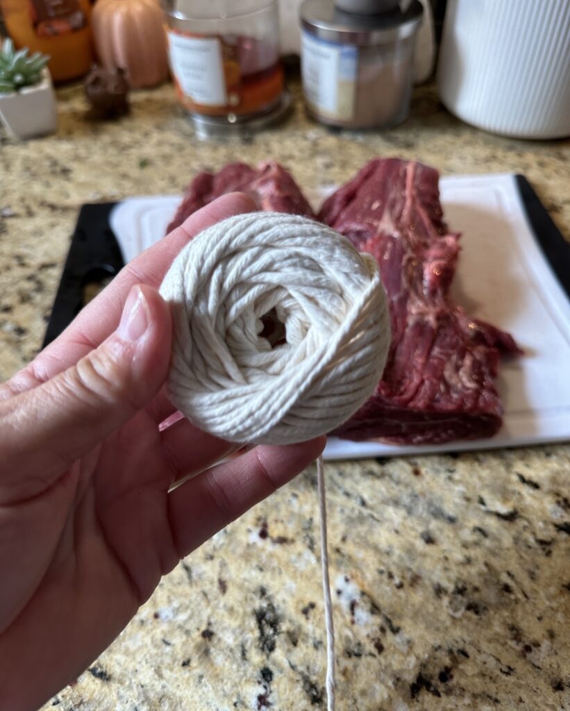 Twine being held in the foreground as two pieces of raw beef tenderloin sit on a cutting board in the background