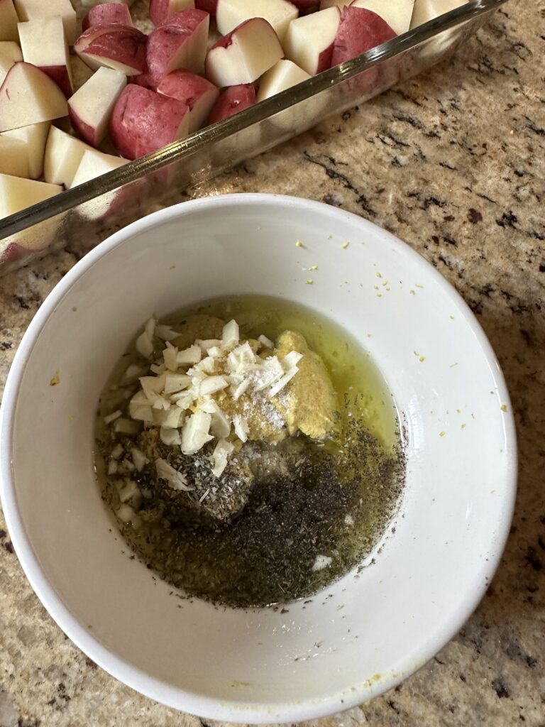 Raw, sliced potatoes in a baking dish next to a bowl with different ingredients for the herb poupon mixture