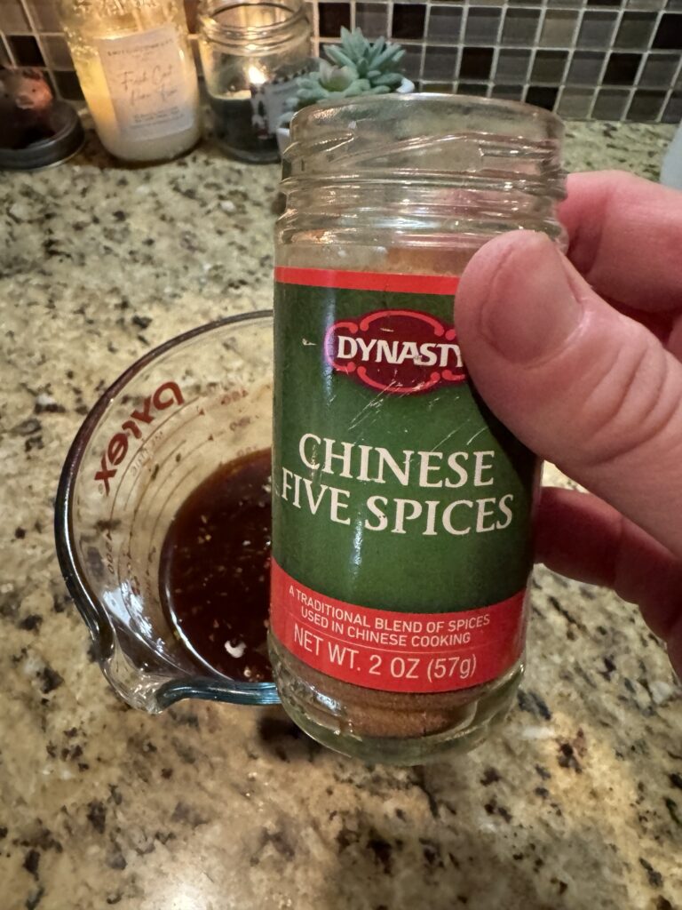 A container of Dynasty brand Chinese five spice being held over sauce in a measuring cup