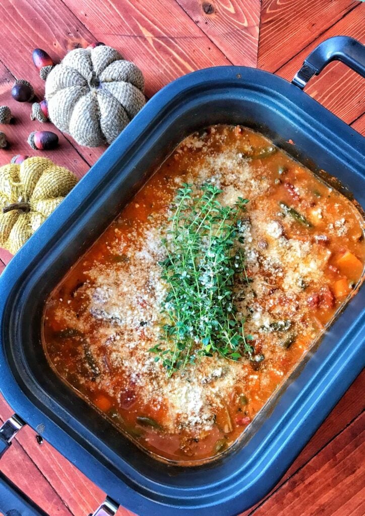 A slow cooker with a rich brownish soup broth and vegtables like carrots and beans