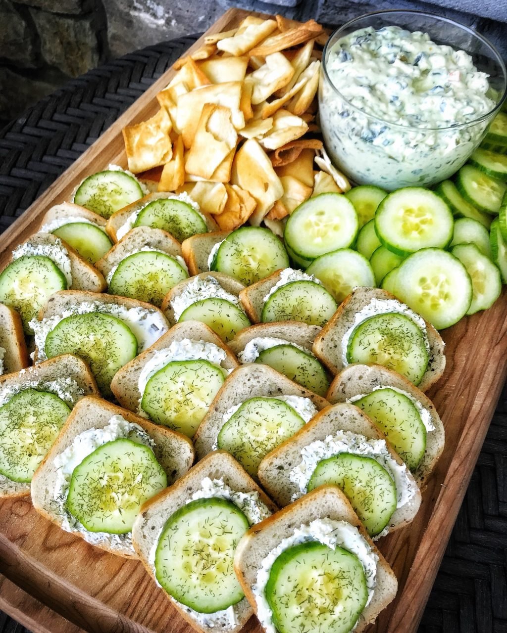 Rye toasts with cream cheese and cucumber slices on a cutting board with a white dip in a glass container and pita chips