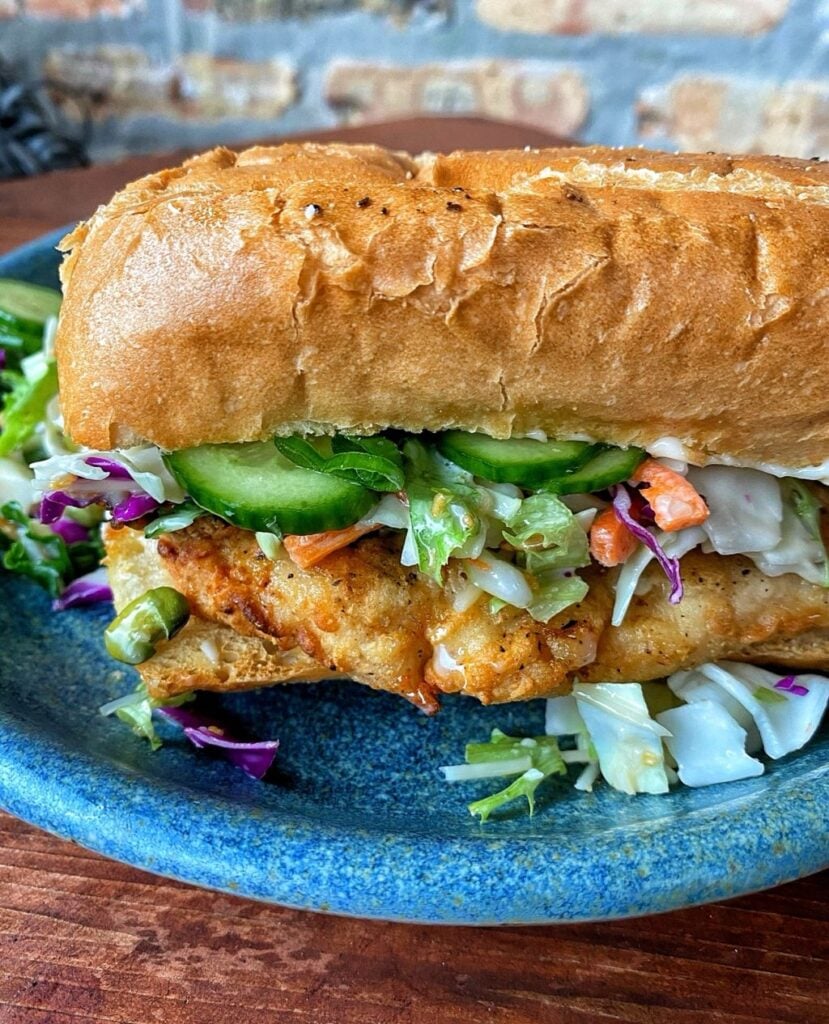 a chicken tender fried and layered on a hoagie roll with curry mayo and a crunchy slaw with marinated cucumbers and displayed on a blue clay plate