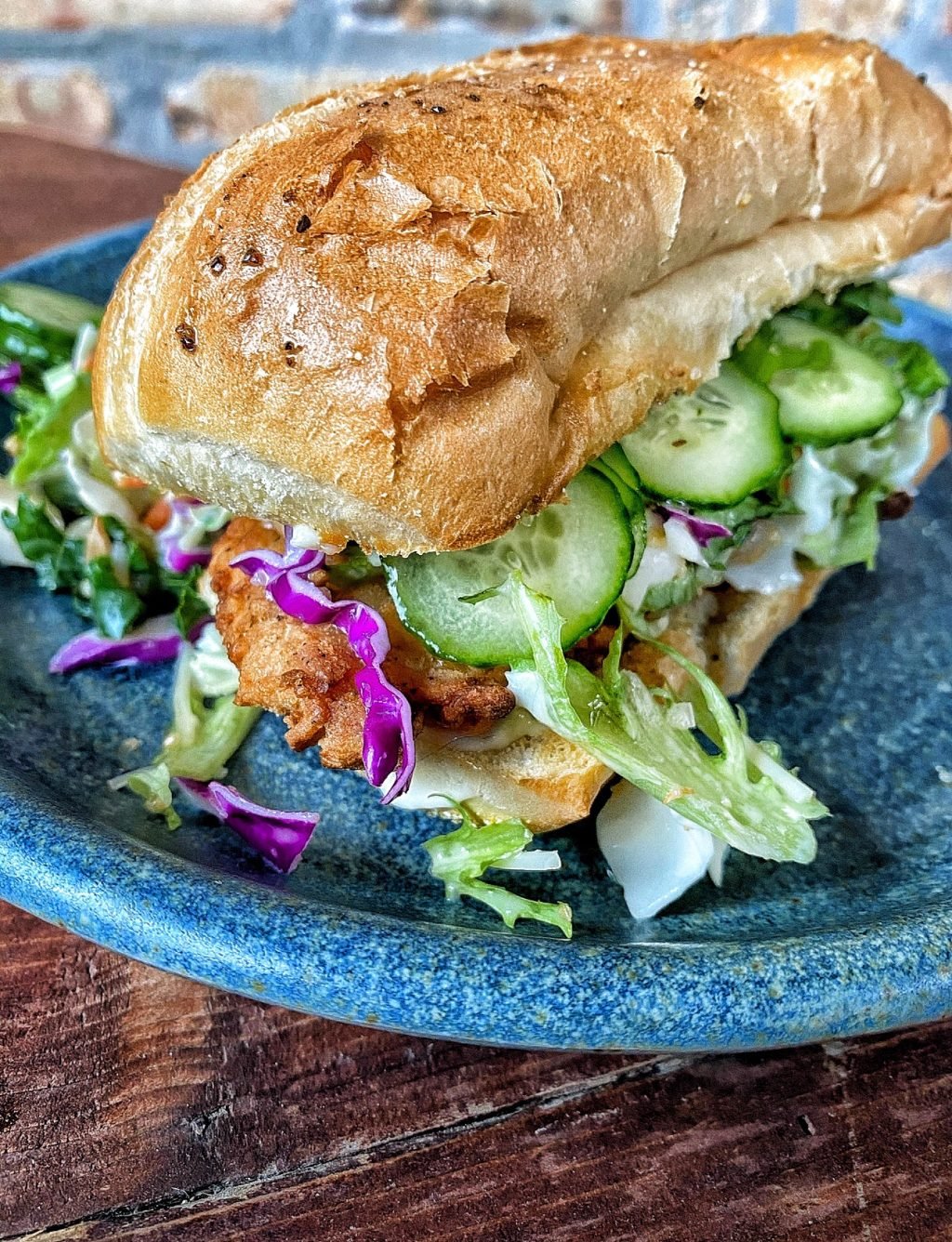 The finished chicken sun sandwich. A chicken tender topped with salad and cucumbers sits inside a split hoagie roll on a blue plate