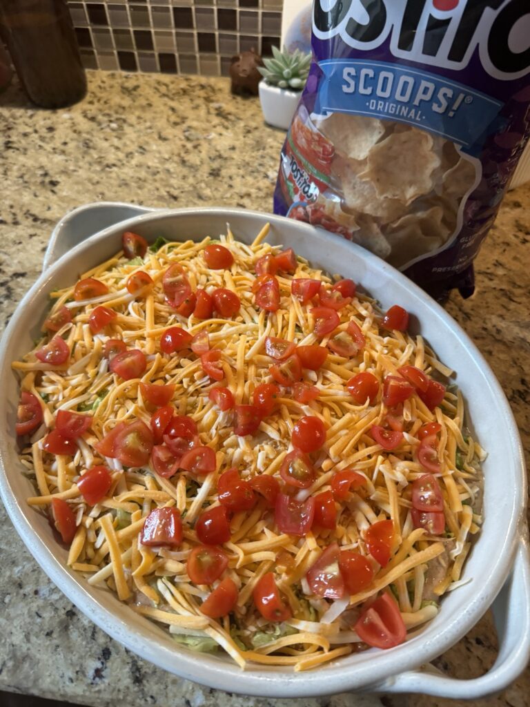 Sliced tomatoes over the dip, all of which is in a baking dish next to a bag of Tostitos Scoops chips