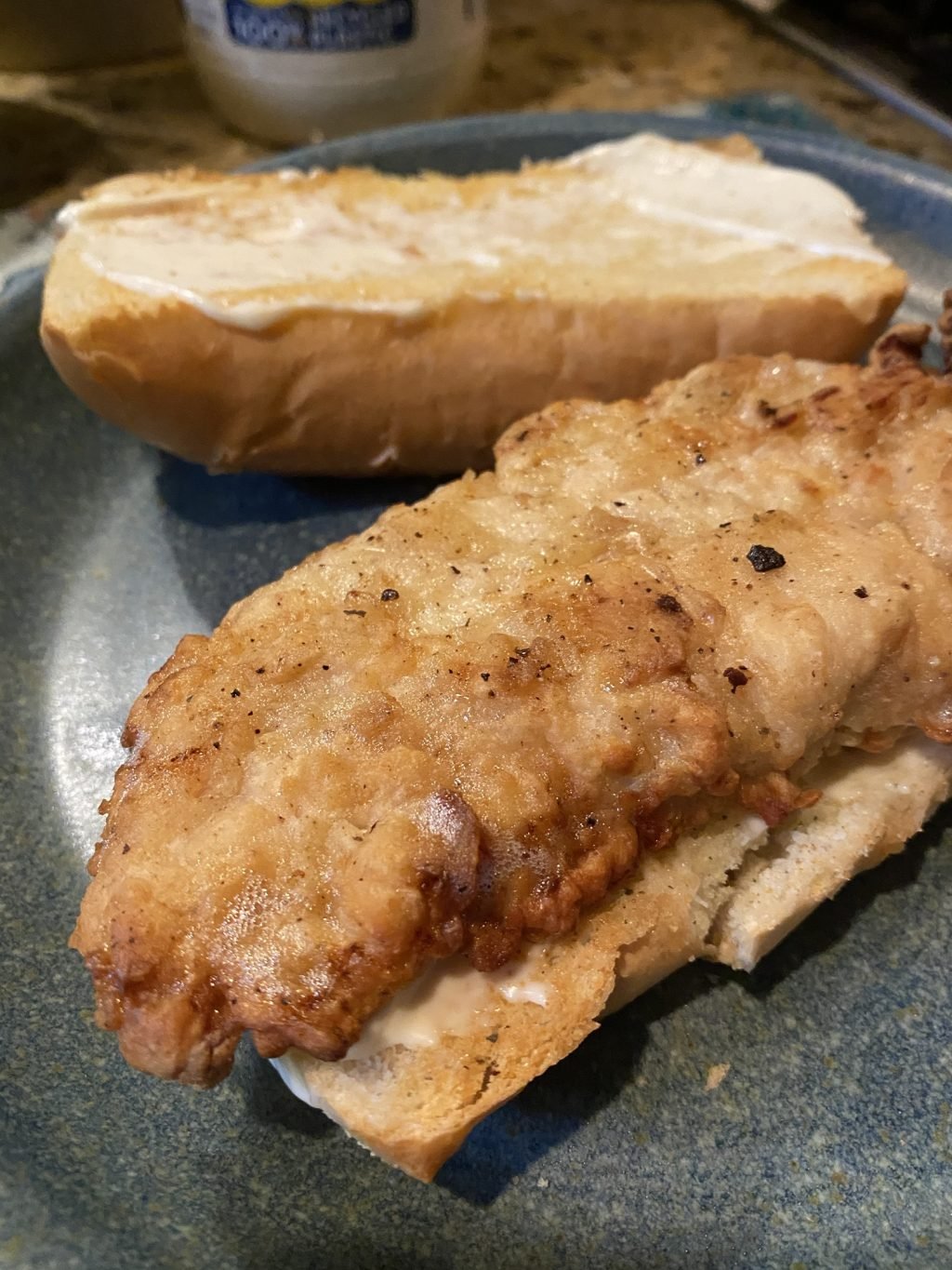 A chicken tender on an open faced hoagie roll. The other half of the roll is behind the tender and has mayo spread on it