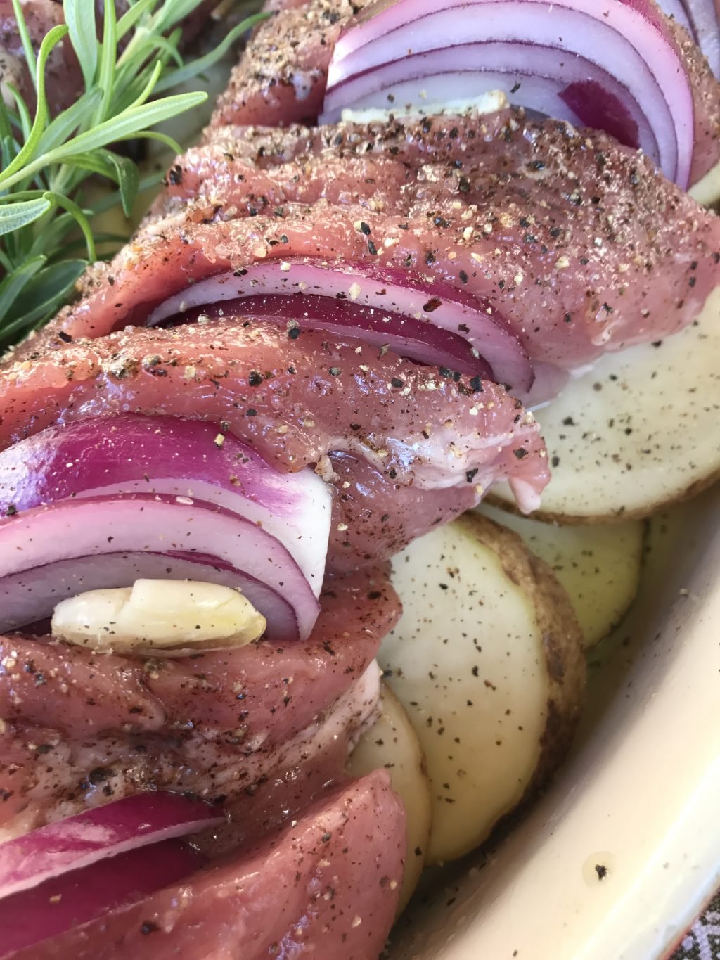 A close up of sliced, raw pork with onions and garlic stuffed into it. It is on a bed of raw potatoes