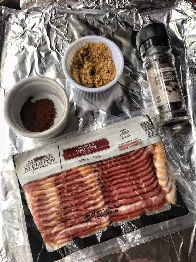 A package of raw bacon on foil in front of containers of cinnamon, brown sugar, and pepper