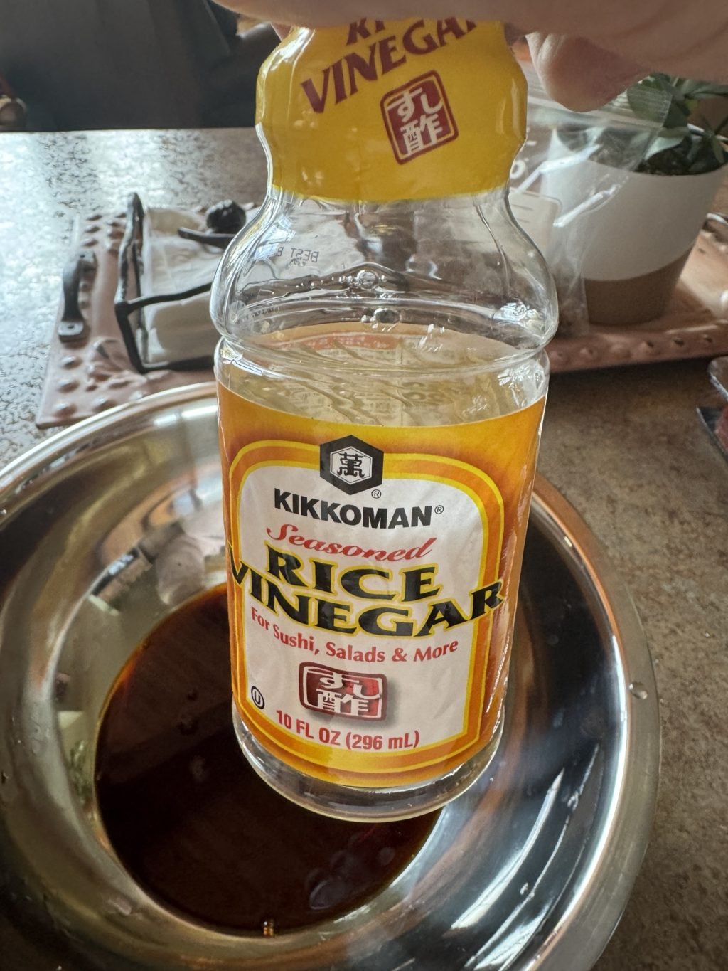 An empty glass bottle of Kikkoman seasoned rice vinegar being held over a metal bowl of its former contents