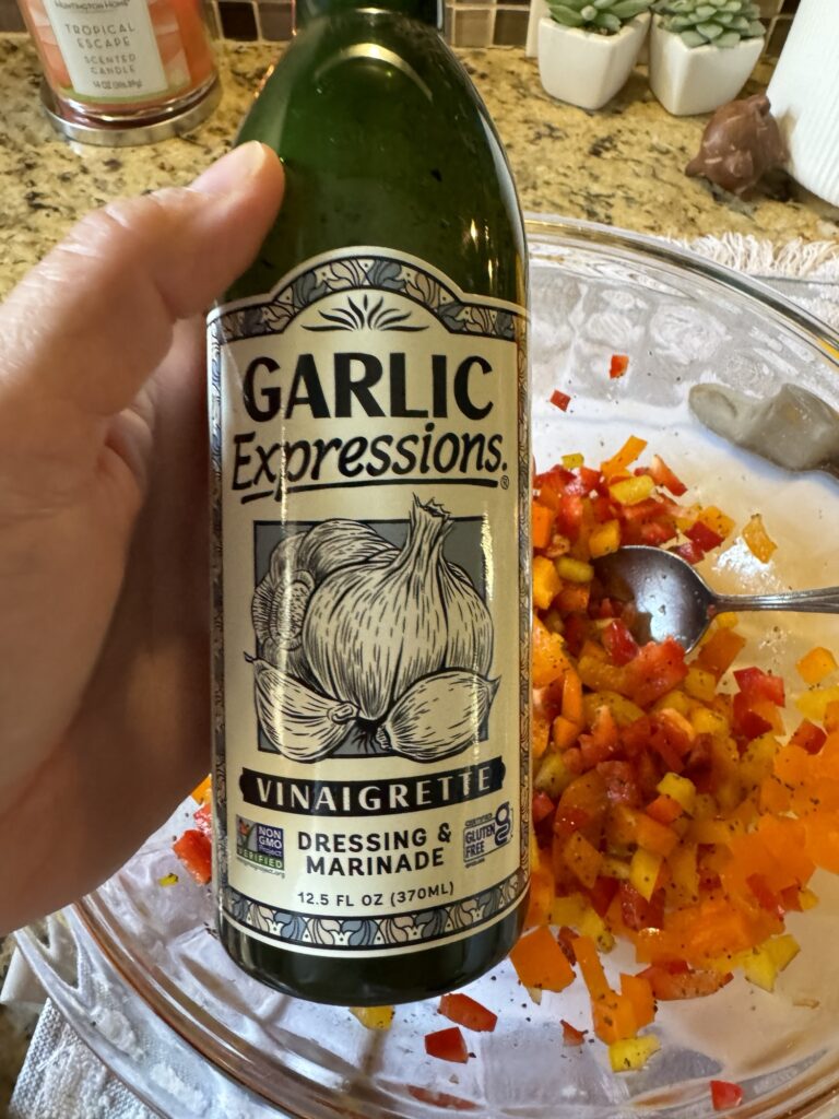 A bottle of Garlic Expressions vinaigrette held over a glass bowl of chopped orange, yellow, and red peppers