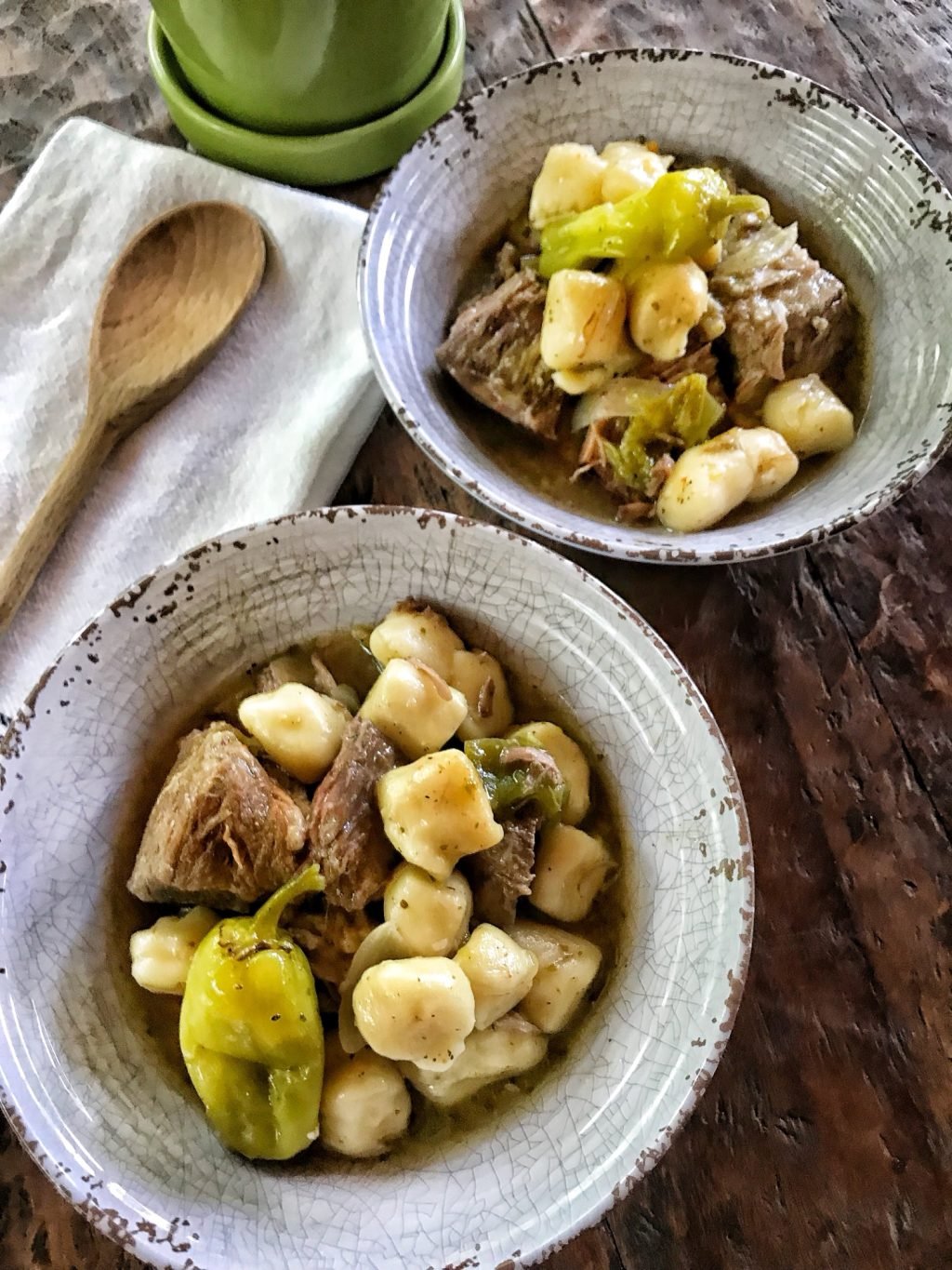 Two bowls of Italian Beef stew with potato dumplings and peperoncini peppers next to a wooden spoon on a kitchen towel