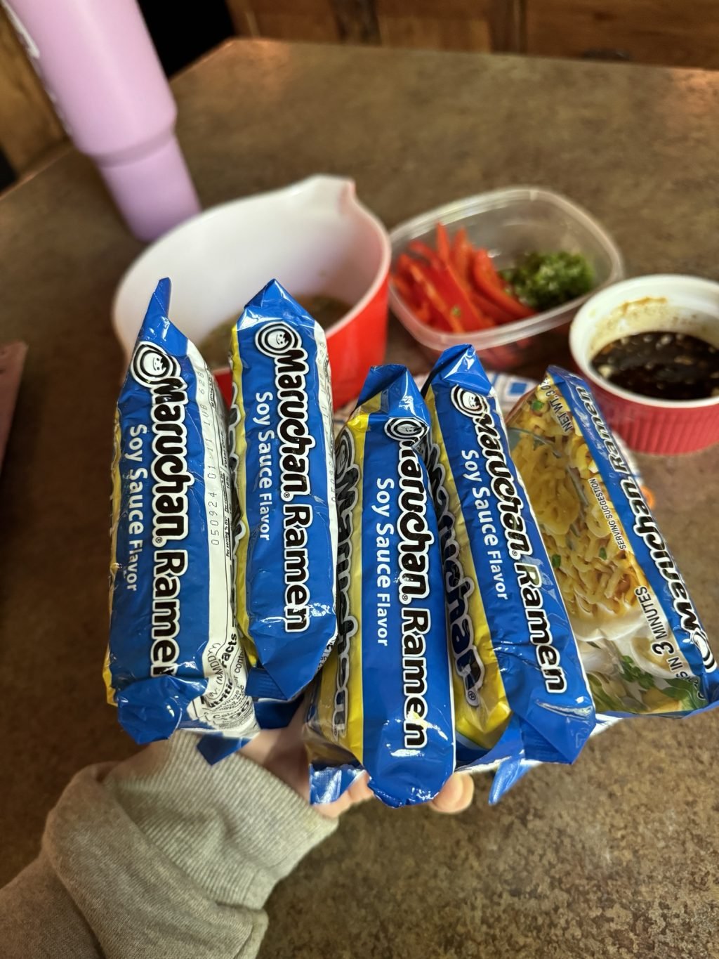 Five packets of Soy Sauce flavored Maruchan Ramen being held over the other ingredients for the cold ramen salad.