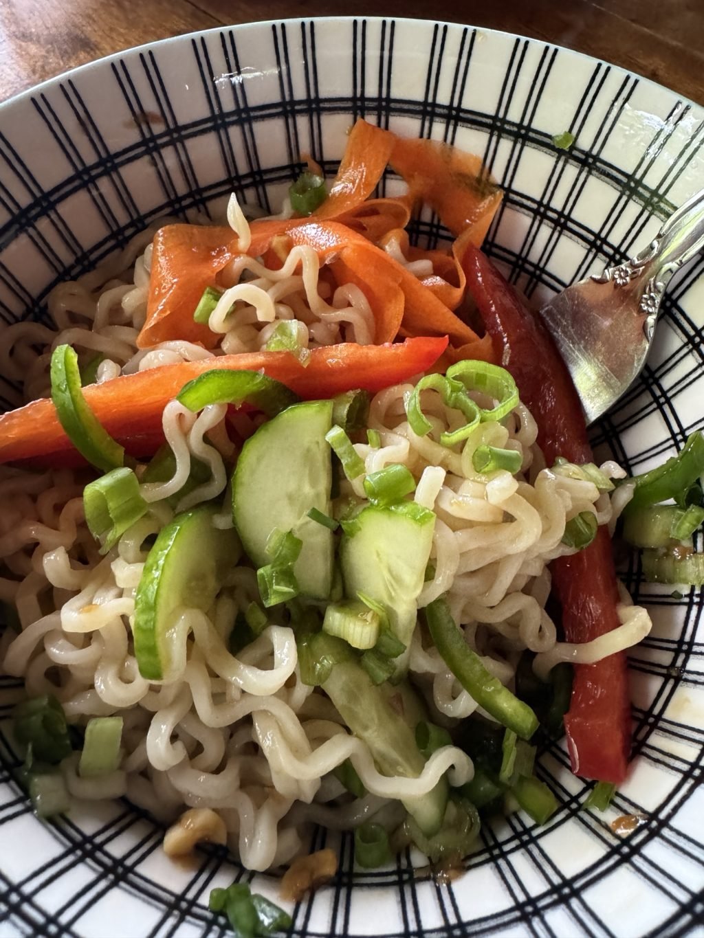 Cold ramen noodles mixed with sliced red bell peppers, cucumbers, and green onion with a fork in a bowl.